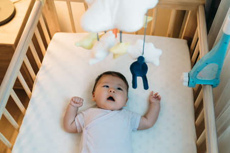 high angle view lovely asian newborn girl lying in the baby bed is staring at the hanging toy with...