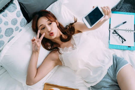 high angle shot sexy asian female lying on bed is making v sign and pursing lips while having fun...