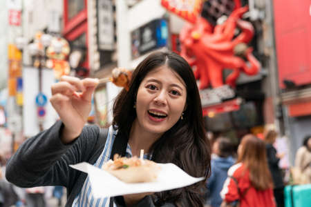 smiling Asian female sight-seer holding showing a takoyaki ball on pick and looking at camera while visiting shinsaibashi suji and doutonbori downtown Osaka japanの写真素材