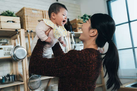 dutch angle shot joyful asian new mother is smiling and lifting her cute baby while playing with her in a bright kitchen at home.の写真素材