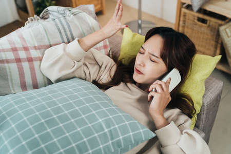 closeup angry confused asian female is having an unpleasant talk on the phone with a raised hand gesture while lying on the couch in the living room at home.の写真素材