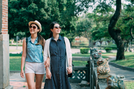 two cheerful asian sister besties are looking around and enjoying the nature while walking arm in arm in a garden with stone lion statues outdoors in summer.の写真素材