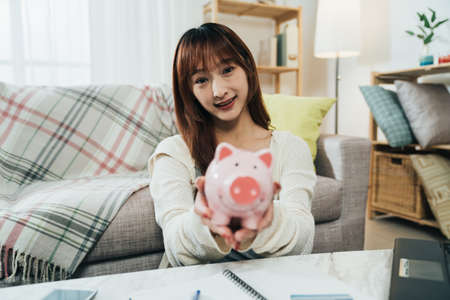 selective focus portrait of a cute asian girl smiling at the camera is giving showing a piggy bank in hands with extended arms in a cozy living room at home.の写真素材