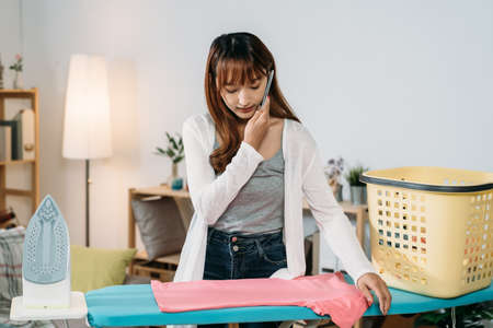 front portrait pretty asian young wife is talking on the phone while standing by the board ironing clothes in a cozy living room at home.の写真素材