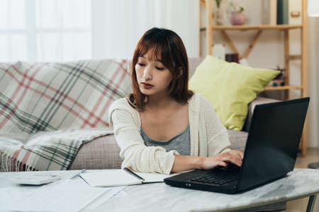 portrait beautiful asian female is looking at the financial document and entering information on the laptop while filing tax online in the living room at home.の写真素材