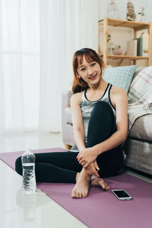 vertical charming asian girl in sportswear is smiling at camera while taking a rest on the yoga mat with her phone and a bottled water after exercise at home.の写真素材
