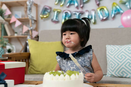 lovely asian toddler baby holding cake knife and licking delicious cream off her mouth at birthday party in a decorated home living roomの写真素材
