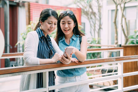 asian woman is pointing on the screen of her friendâs smartphone as they are having fun looking at pictures by a railing at a cozy outdoor area in summer.の写真素材