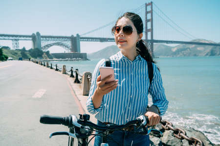 happy Korean woman is using navigation app on smartphone while walking with a bike along the road on a sunny holiday by the ocean in San Francisco California.の写真素材