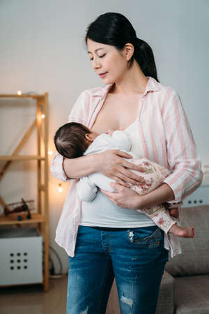 portrait affectionate asian mother is gazing at her infant child in arms while breastfeeding in a standing position at home.の写真素材