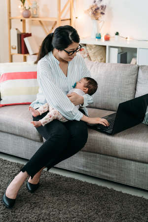 vertical shot busy asian working mother is holding her sleeping baby while using computer on the couch in the living room at home.の写真素材