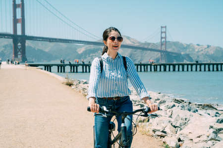 carefree japanese girl wearing sunglasses is riding bicycle along the beach on summer holiday with...