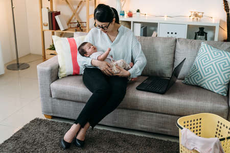 asian businesswoman mother is consoling and putting her baby daughter to sleep while working from home on the couch in the living room at home.の写真素材