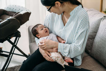 lovely asian baby girl lying in her smiling career momâs arms is looking up with innocence on the couch at home.の写真素材