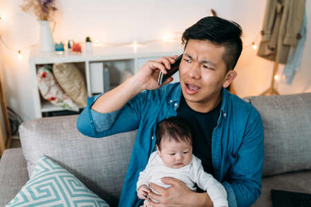 annoyed asian dad holding a smpartphone is having an unpleasant conversation while taking care of his baby daughter on the couch in the living room at home.の写真素材