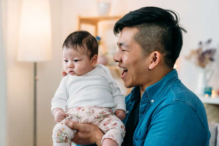 closeup playful asian daddy is holding and smiling at his innocent tiny new baby in hands at a cozy home living room background.の写真素材