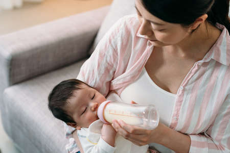 high angle shot affectionate asian mother is looking at her newborn girl drinking milk from the bottle in her arms on the sofa in the living room at home.の写真素材