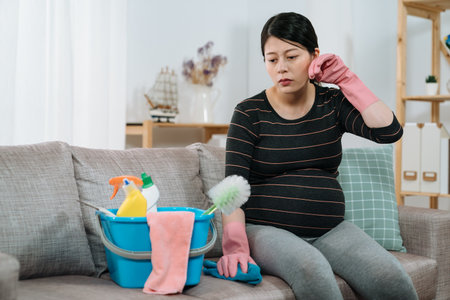 uncomfortable asian expectant pregnant mom is mopping sweat with hand while taking a rest on sofa in the living room after doing housework on a overheating summer day.の写真素材