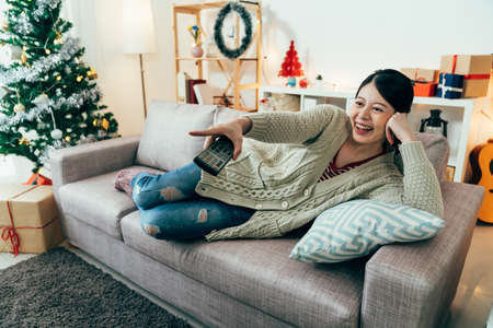 asian female couch potato is pointing at screen while watching funny tv shows. woman lying on sofa on her side is having a hearty laugh. indoor recreation ideas for Christmas holiday. real momentsの写真素材