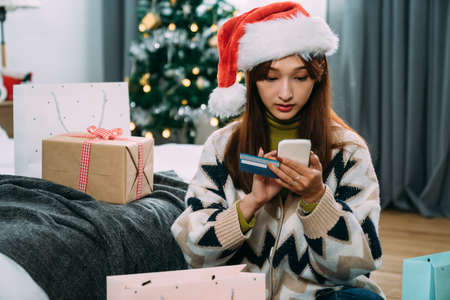 asian female wearing a Christmas hat is entering credit card information on the phone while shopping online and paying for gifts in a bright festive bedroom.の写真素材