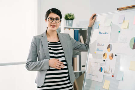 portrait of a smiling asian expectant businesswoman looking at the camera while keeping a hand on the white board and anther on belly in bright creative officeの写真素材