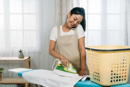 carefree asian wife wearing an apron is holding mobile phone on her shoulder talking and ironing clothes in a cozy bright laundry room at home with daylight.の写真素材