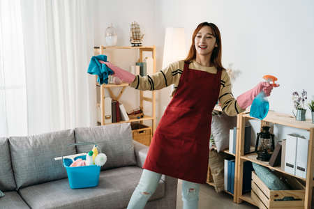 happy asian woman wearing an apron holding a rag and cleanser spray bottle is dancing while cleaning house in the living room at home.の写真素材