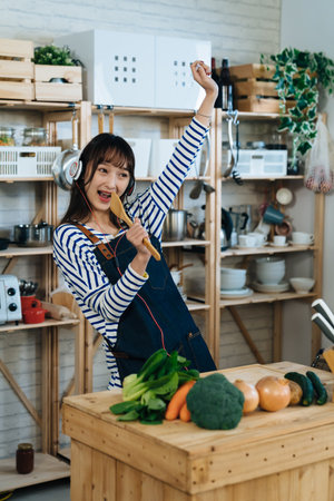 vertical shot of an energetic asian wife listening to music from headphones and having fun singing into the spatula with a raised arm in the kitchen at home.の写真素材