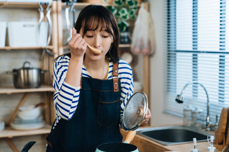 closeup of an Asian young wife is tasting the soup from spoon with a lid in hand while cooking during daytime in the kitchen at home.の写真素材