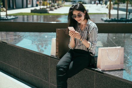 Asian trendy female is enjoying social media feeds on the smartphone while sitting near a fountain for a break from shopping in the city on a sunny day.の写真素材