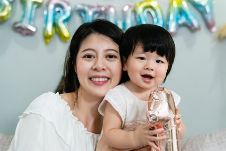 closeup portrait of beautiful asian mother and lovely baby girl smiling at camera with their heads close at blurred background with colorful birthday decorationの写真素材