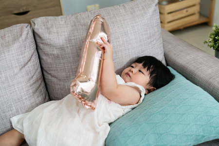 chubby asian one year old baby girl lying on the sofa cushion and playing with a golden number...