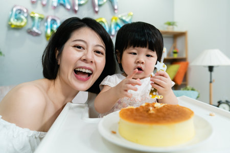 closeup portrait of a smiling asian mother facing camera and posing close to her innocent baby girl who is playing with the candle on cake at birthday partyの写真素材