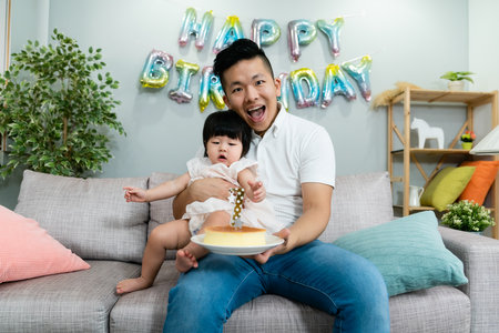 happy asian dad sitting on sofa is smiling at camera with a cake in hands and his baby daughter in...