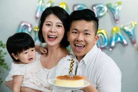 joyful asian father and mother celebrating first birthday of their baby daughter with a cake while smiling at the camera at home with festive decorationの写真素材