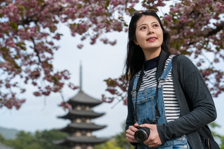 cheerful asian Japanese girl photographer looking into distance at beautiful landscape with Kofuku-jiÃ¢ÂÂs gojunoto pagoda and cherry blossom at background in nara japanの写真素材