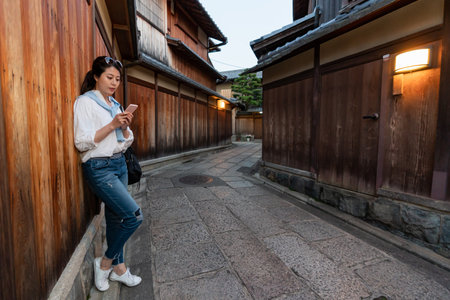 full length of relaxed asian Japanese girl visitor leaning against wooden wall and looking at her cellphone in illuminated Ishibeikoji alley in gion Kyoto japan at sunsetの写真素材