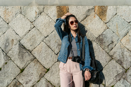 stylish asian Japanese woman tourist wearing sunglasses posing against a rock wall in uji shi Kyoto japan. she pushes her hair back while looking away into space in the sunの写真素材