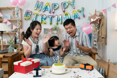 innocent asian baby girl looking at her father while singing songs before blowing out candle at birthday party in a cozy home interior with colorful decorationの写真素材