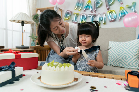 smiling asian mother wiping baby girl's face with a tissue as she is playing with a cake server at the birthday party at homeの写真素材