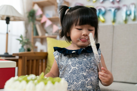closeup of happy asian toddler baby girl licking yummy whipped cream on face and looking playing with the cake knife on her second birthday party at homeの写真素材