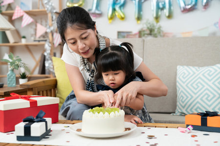 loving asian mother holding toddler baby girl's hands while cutting delicious birthday cake together with a knife on the anniversary celebration at homeの写真素材