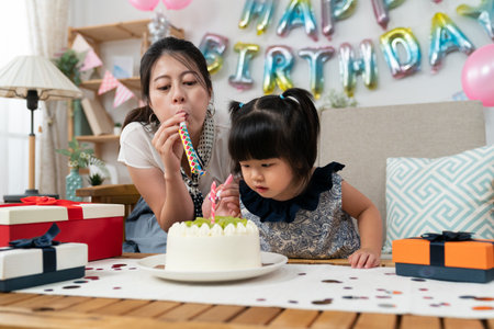 playful asian mother blowing party horns as her cute baby daughter is staring at the delicious birthday cake with curiosity in the birthday party at homeの写真素材