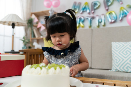 closeup view of innocent asian baby girl using knife to cut cake while celebrating her second birthday at home with colorful party decorationの写真素材