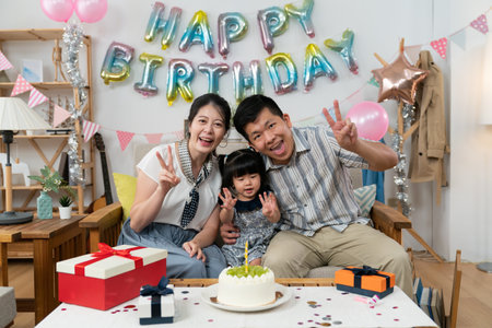 happy asian small family looking at camera with victory hand gestures at the baby girl's second birthday celebration party at home with pink balloon and garlandの写真素材