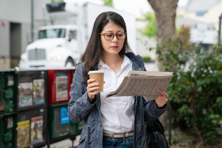 asian chinese business woman carrying morning coffee and learning about current events on papers while walking away from newspaper box on her way to work in palo alto city California usaの写真素材