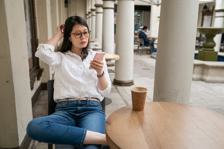 leisure asian business lady wearing earphones binge watching online tv shows on phone during lunch break at cafe in palo alto city California usa. she folds legs and scratches head while watchingの写真素材