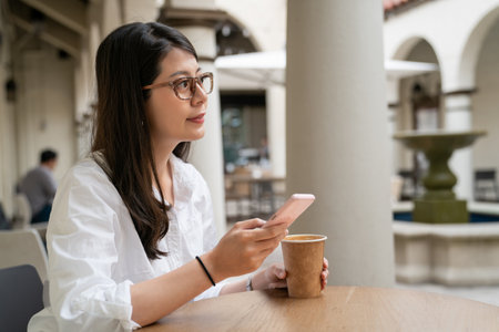 pretty asian chinese business lady looking into distance in thought while sending sms text message on phone to client at outdoor seating of cafe in palo alto city California usaの写真素材