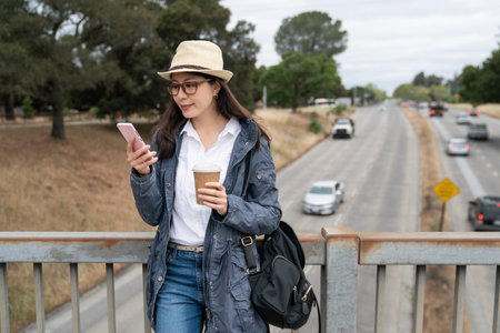 happy asian Korean female backpacker holding coffee and enjoying social media feeds on phone while leaning relaxing against bridge railing above a public road in palo alto city California usaの写真素材