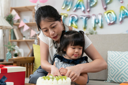 cute naughty Korean baby girl sticking her tongue out as her mother is dividing birthday cake and holding her hand to keep it off from the dessert at homeの写真素材